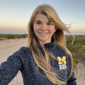 Mariah Schlis-Elias outside smiling at camera with a dirt road and sunset behind her.